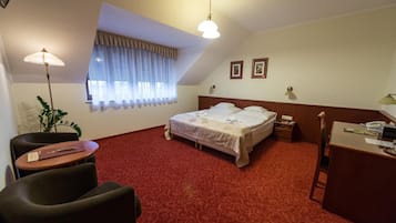 A hotel room with a bed, a desk, a chair, a window with curtains, and a plant.