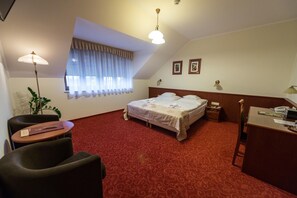 A hotel room with a bed, a desk, a chair, a window with curtains, and a plant.
