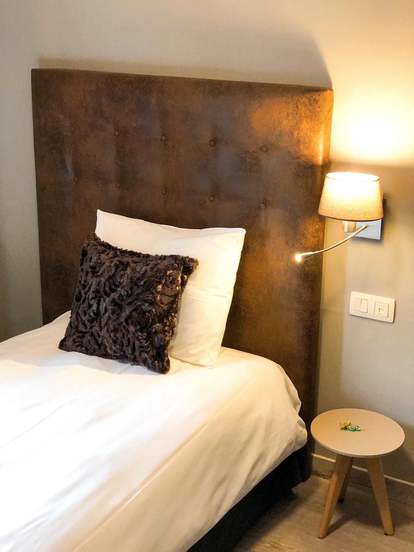 A bed with white linens and a dark patterned pillow, a wall-mounted lamp, a small round table with a stool, and a power outlet.
