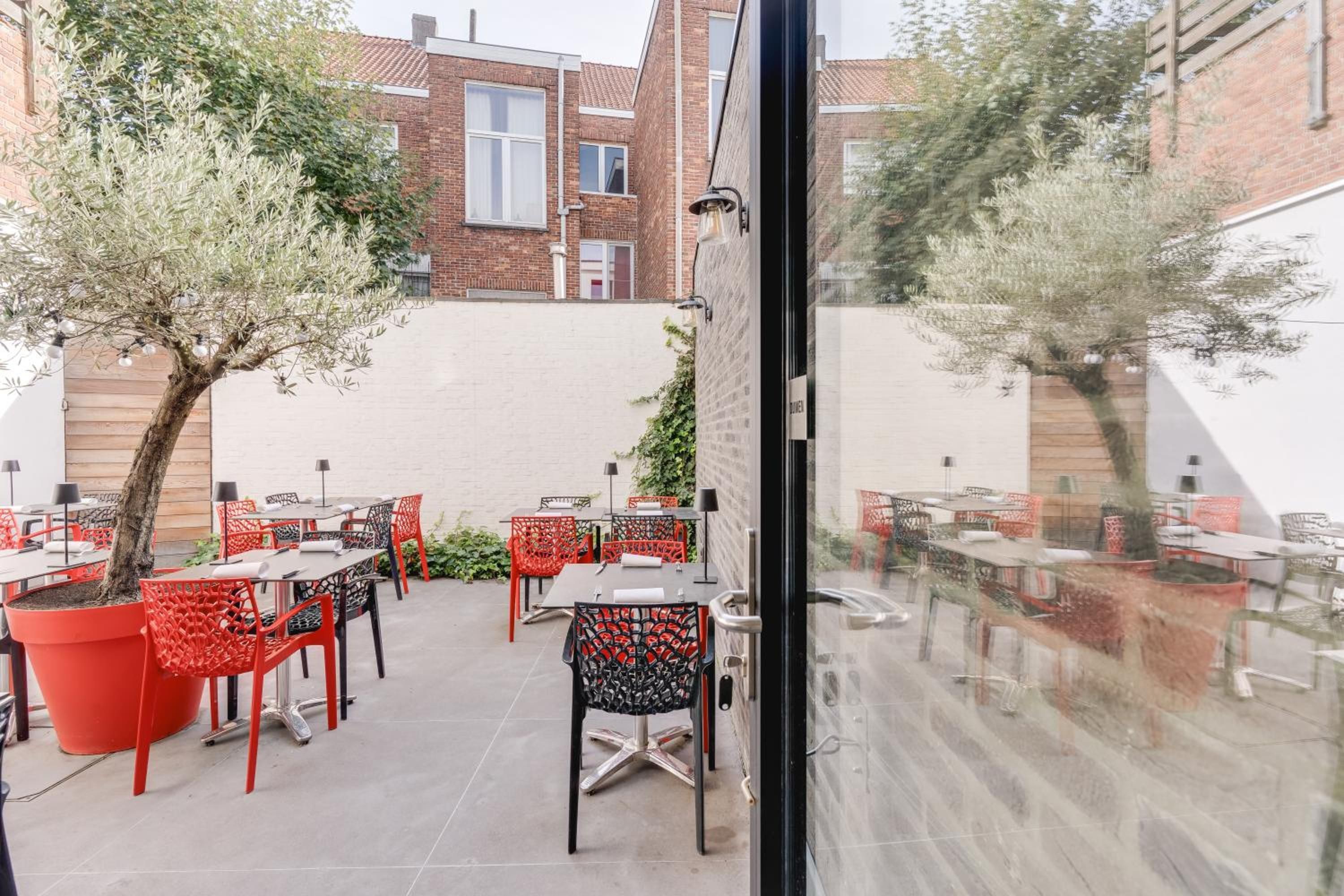 Une terrasse extérieure avec des tables et des chaises, un bâtiment en briques en arrière-plan et un arbre au premier plan.