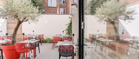An outdoor patio with tables and chairs, a brick building in the background, and a tree in the foreground.