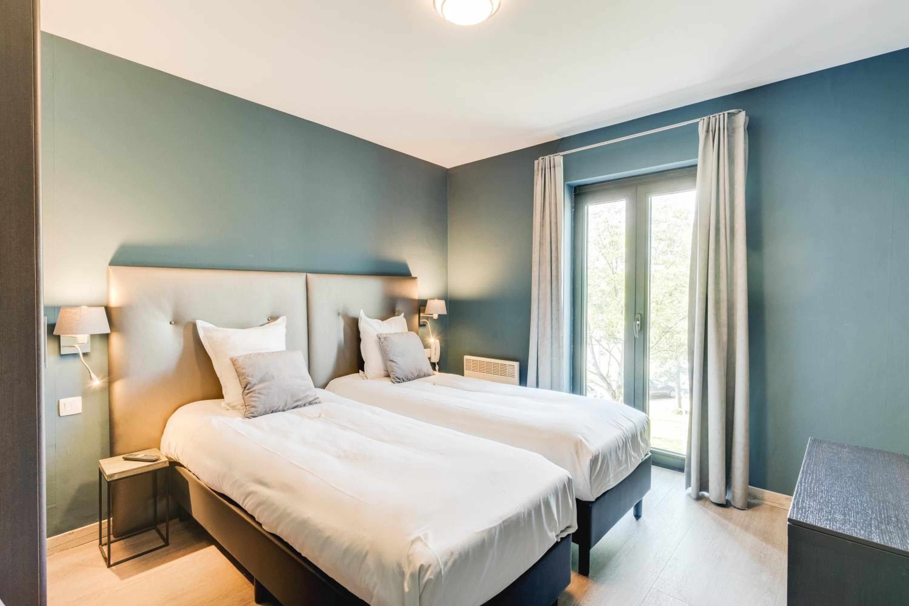 A modern bedroom with two beds, a nightstand, a desk, and a large window with curtains.