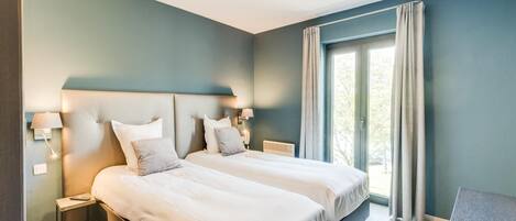 A modern bedroom with two beds, a nightstand, a desk, and a large window with curtains.