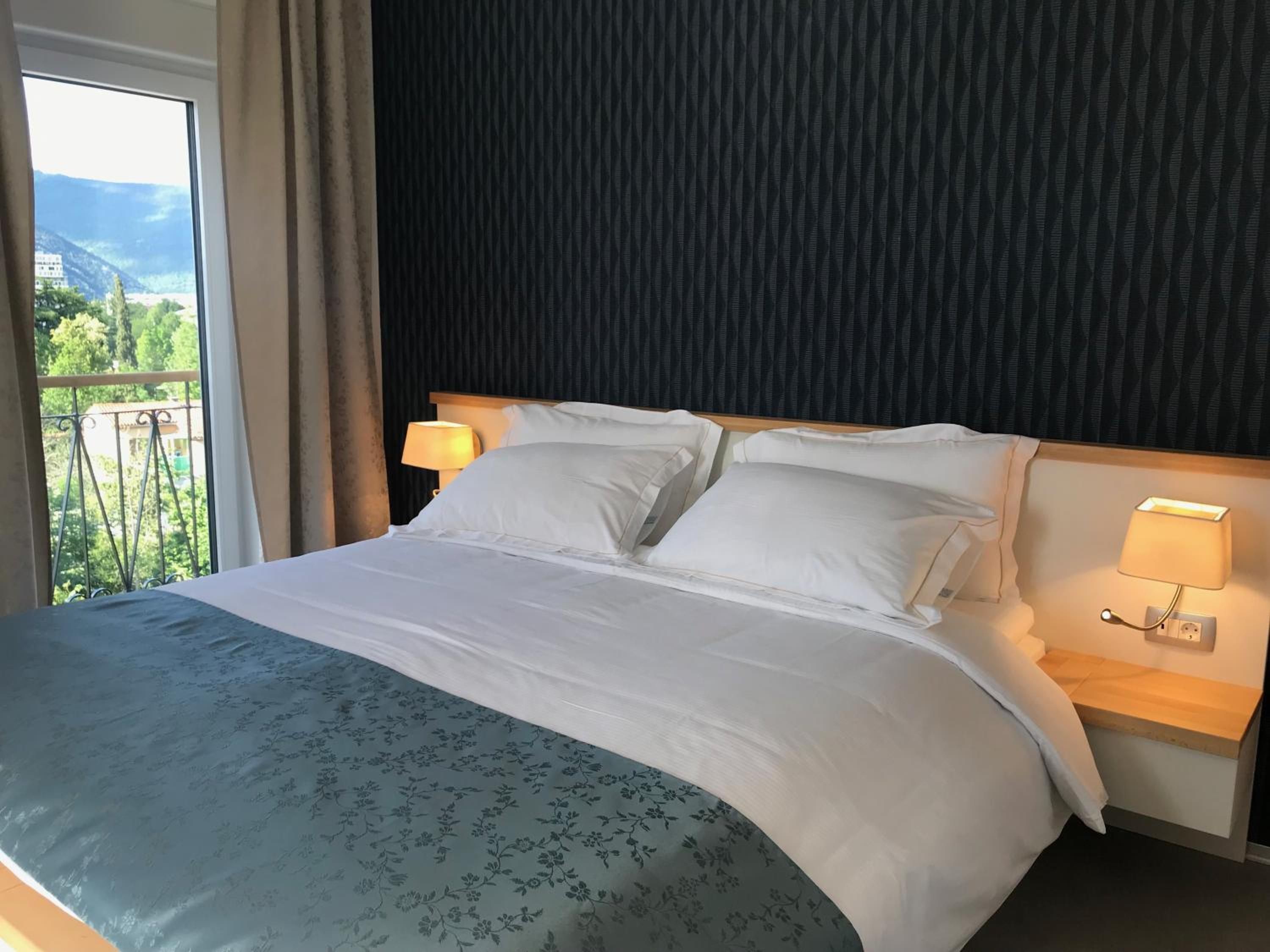 Habitación de hotel con cama, dos lámparas de noche, vistas a la vegetación y montañas a través de una ventana, y una pared oscura con textura.