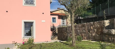 Ein pinkfarbenes Gebäude mit zwei Fenstern, einer Steinmauer und einem Baum im Vordergrund.