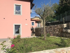 Un edificio rosa con dos ventanas, un muro de piedra y un árbol en primer plano.