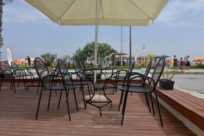 Outdoor seating with tables and chairs, a large umbrella, and a view of the beach. - Korali Hotel (Katerini)
