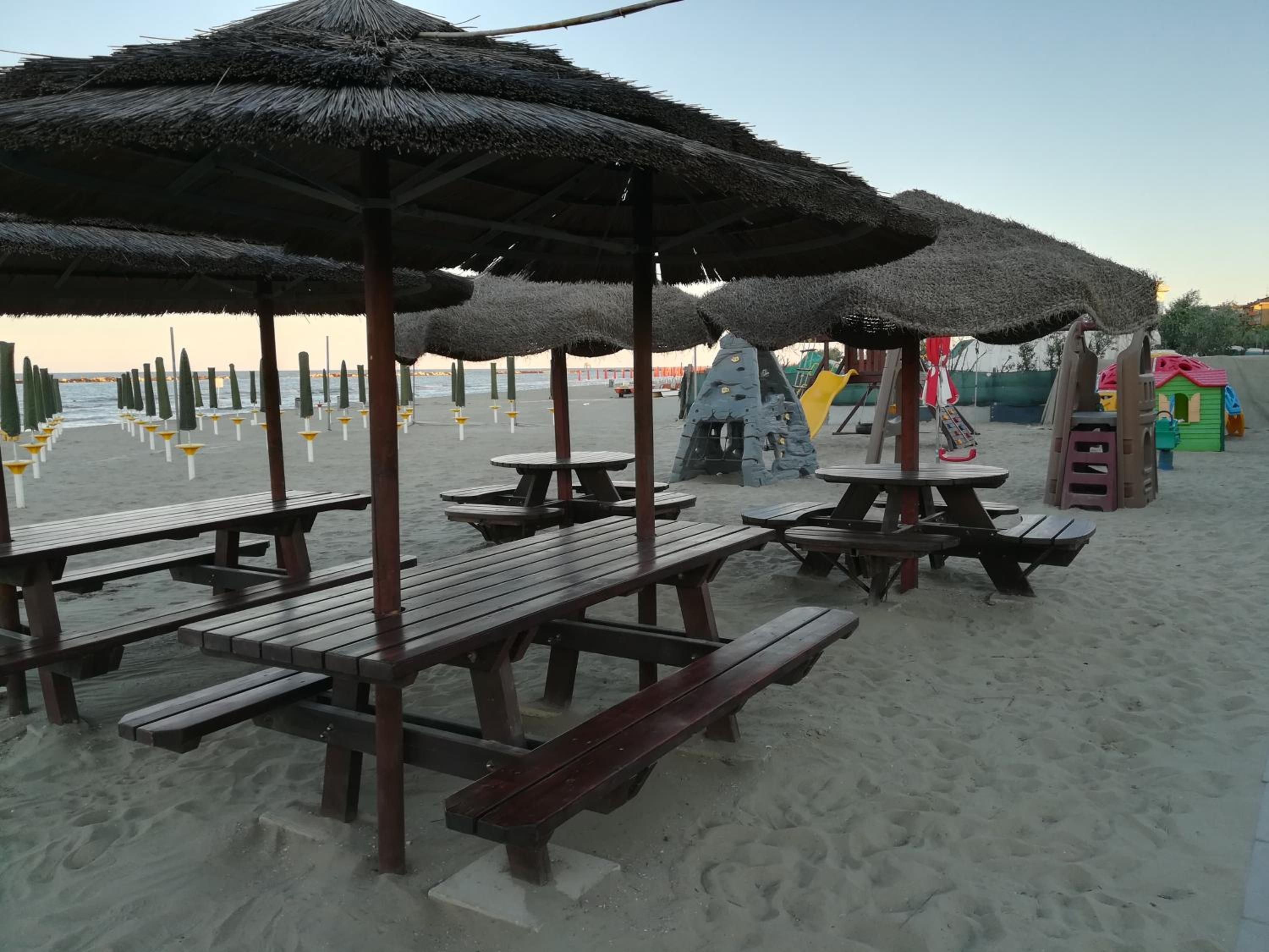Una playa con mesas de picnic y sombrillas, un parque infantil y un cielo despejado.