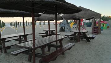 Una playa con mesas de picnic y sombrillas, un parque infantil y un cielo despejado.