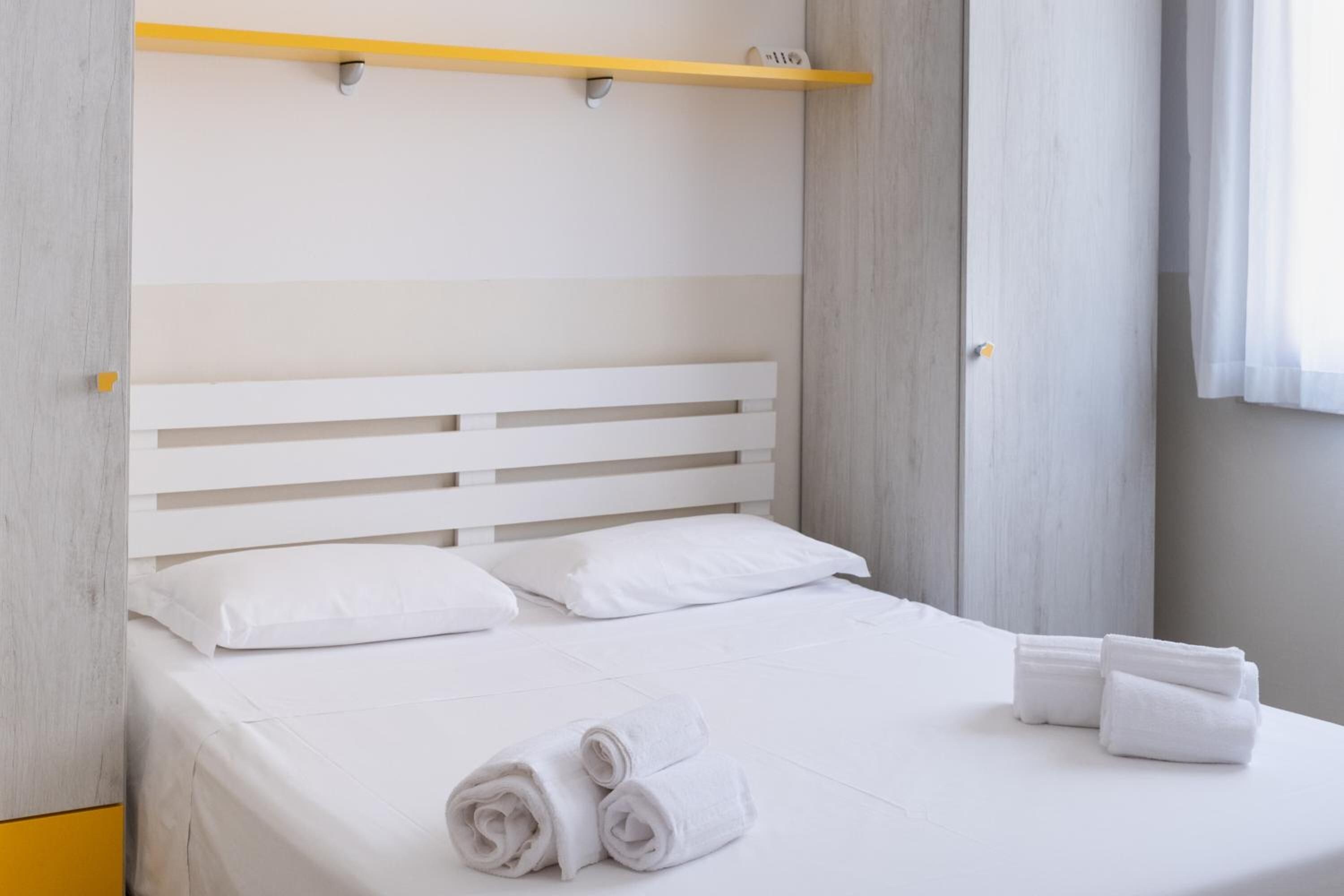 Una cama bien hecha con sábanas blancas y toallas enrolladas, un cabecero sencillo y una pequeña estantería sobre la cama.