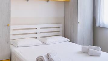Una cama bien hecha con sábanas blancas y toallas enrolladas, un cabecero sencillo y una pequeña estantería sobre la cama.