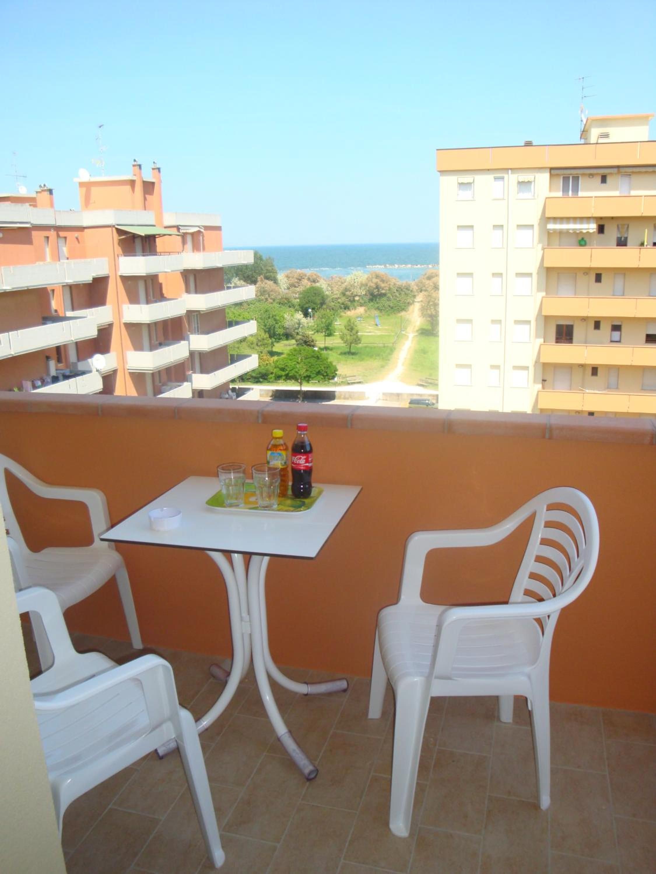A balcony with two white plastic chairs and a small table, overlooking a residential area with a clear view of the sea.