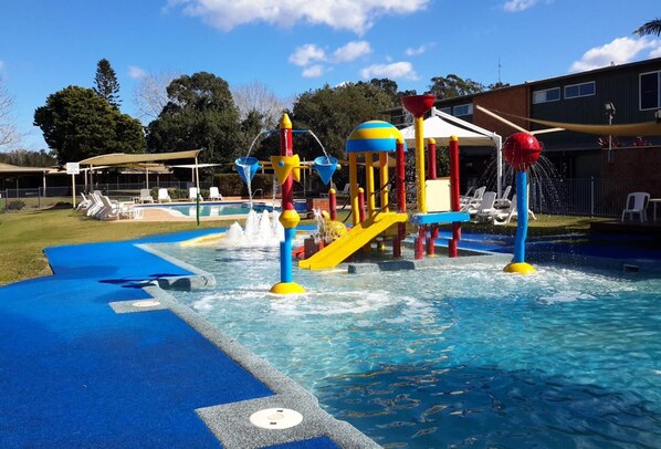 Zona de juegos acuáticos con toboganes y chorros de agua, rodeada por una piscina y tumbonas.