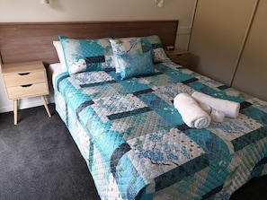 Una cama bien hecha con colcha, una mesita de noche con cajón y una lámpara fijada a la pared.