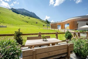 Una terraza de madera con una mesa y bancos, con vistas a una ladera verde y un edificio moderno con amplios ventanales.