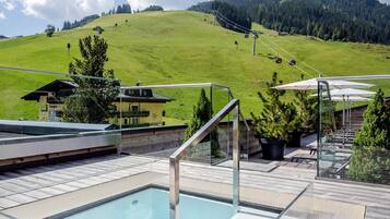 Helderblauwe lucht boven een zwembadcomplex met een glazen pool, een houten terras en uitzicht op bergen.