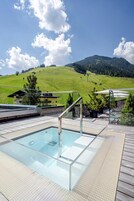 Cielo azul despejado con zona de piscina que incluye una piscina con cerramiento de vidrio, terraza de madera y vistas a la montaña.