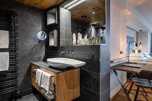 Un baño moderno con un lavamanos independiente, baldosas oscuras, un mueble de tocador de madera, un espejo y una mesita de noche con lámpara.