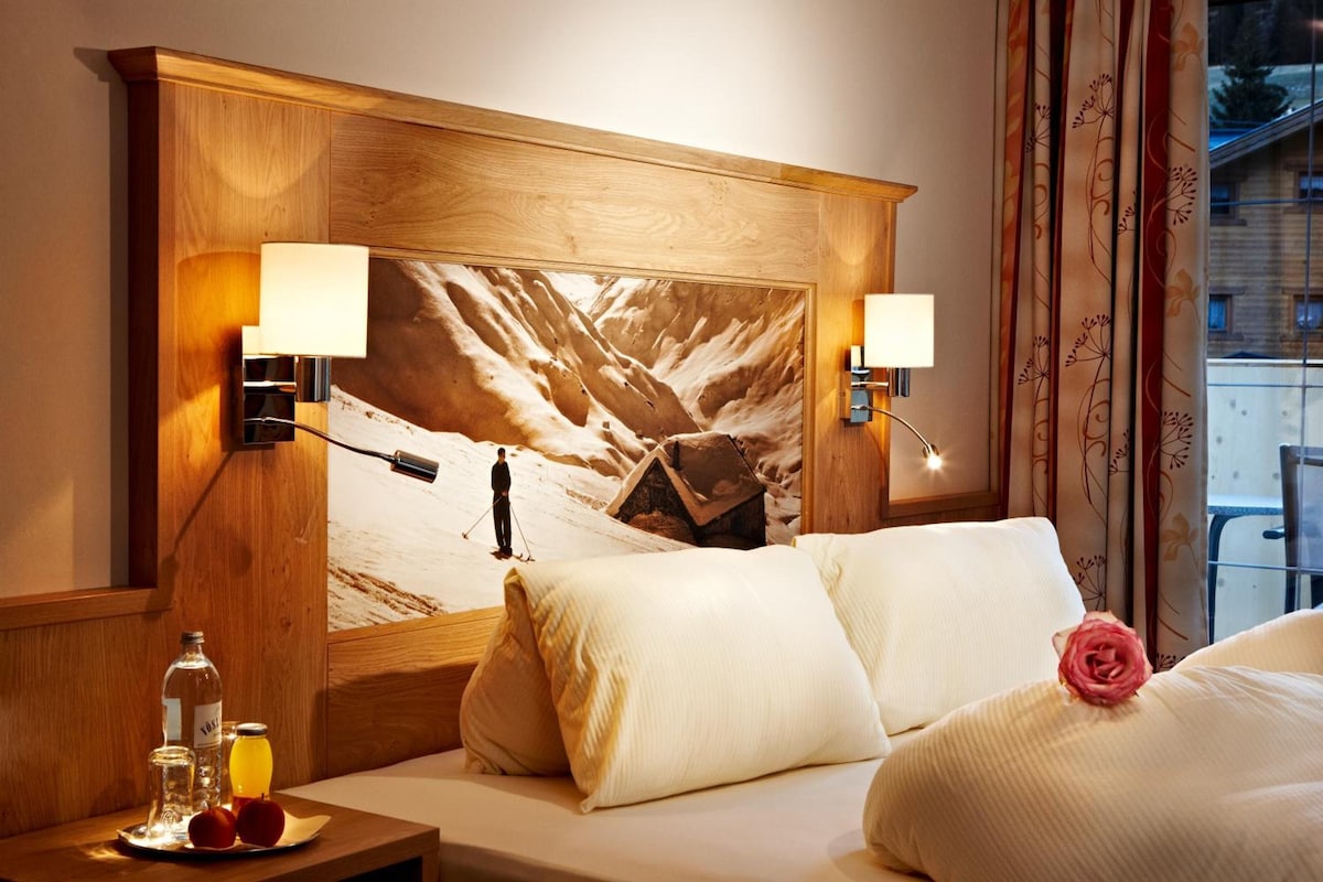 Una camera d'albergo con un letto grande, due lampade da comodino, un comodino con una bottiglia e dei bicchieri, e un quadro raffigurante un paesaggio innevato.