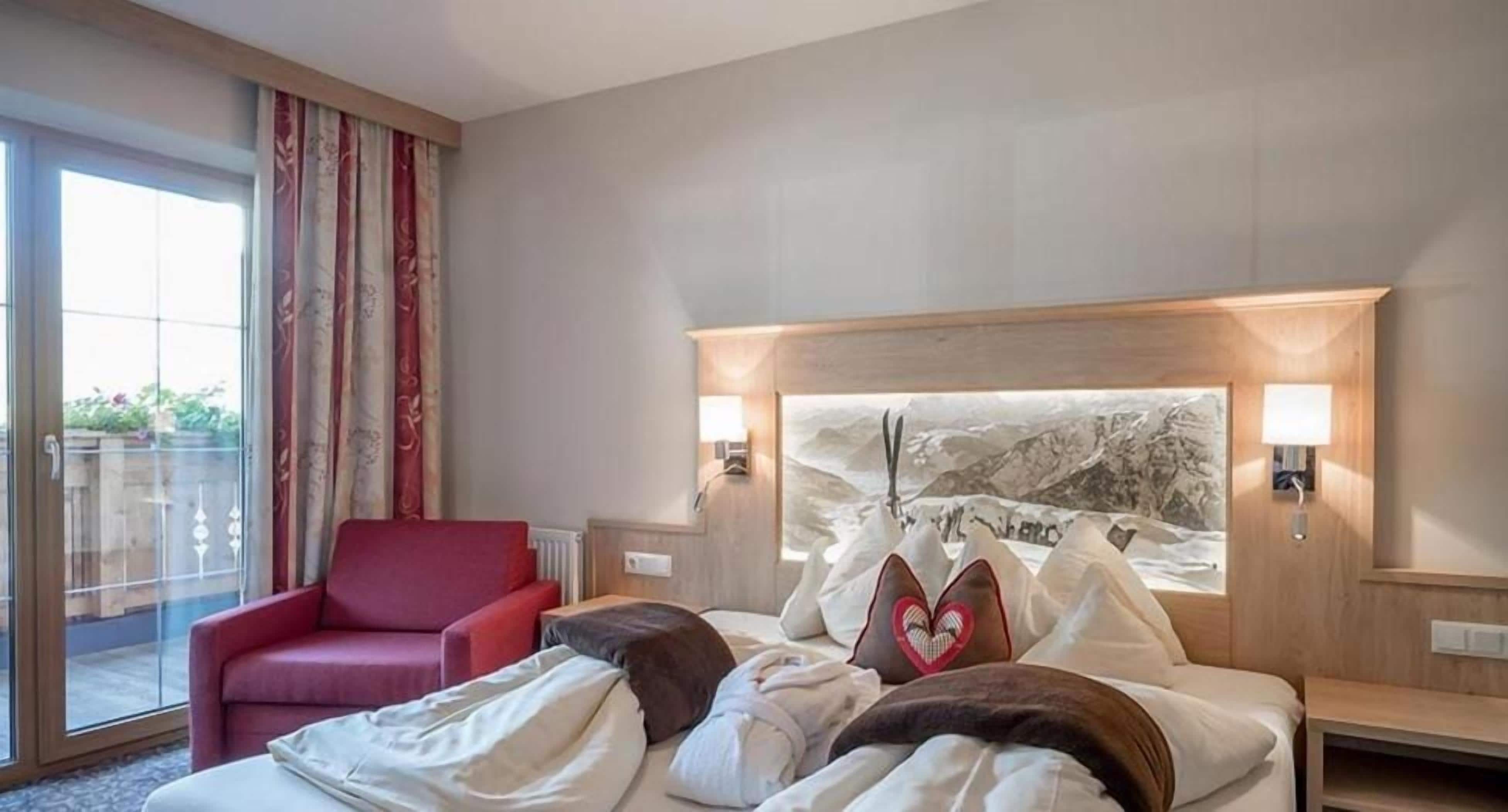 A hotel room with a bed, a red armchair, a large window, and a headboard with a mountain landscape.