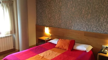 A hotel room with a large bed, a nightstand with a phone, a window with curtains, and a patterned wallpaper.