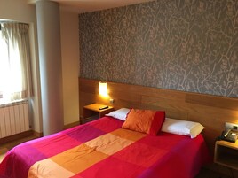 Habitación de hotel con una cama grande, una mesita de noche con teléfono, una ventana con cortinas y un papel tapiz estampado.
