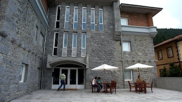 A stone building with a central entrance, two outdoor tables with umbrellas, and a person standing by the entrance.