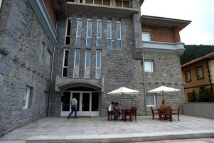 Un edificio de piedra con una entrada central, dos mesas exteriores con sombrillas y una persona de pie junto a la entrada.