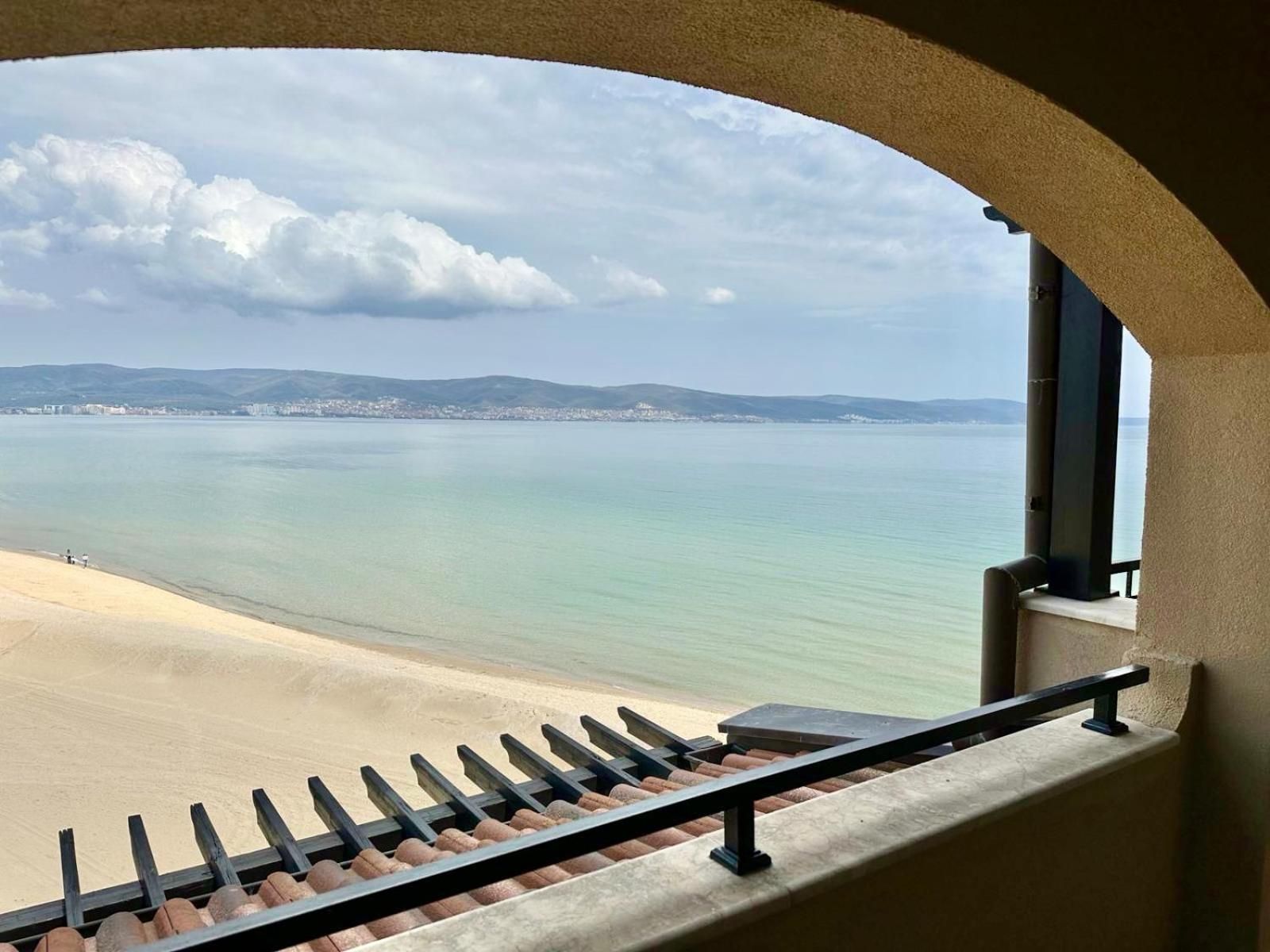 Vista desde un balcón con barandilla, sobre una playa y un paisaje urbano distante.