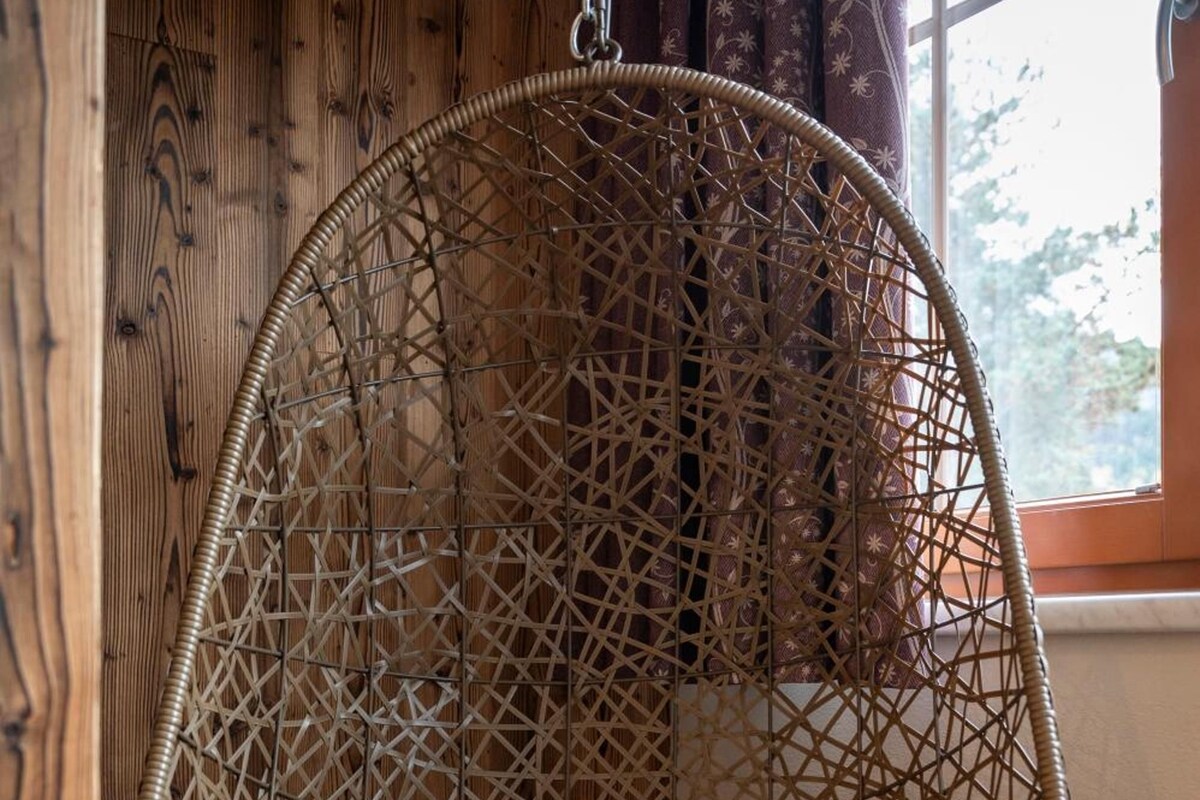 A hanging rattan chair with a cushion, suspended from a chain against a wooden wall with a window in the background.