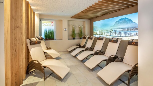 A spa room with multiple sun loungers, a wooden ceiling, a window with a mountain view, and a clock on the wall. - Bliem S Familienhotel (Haus)