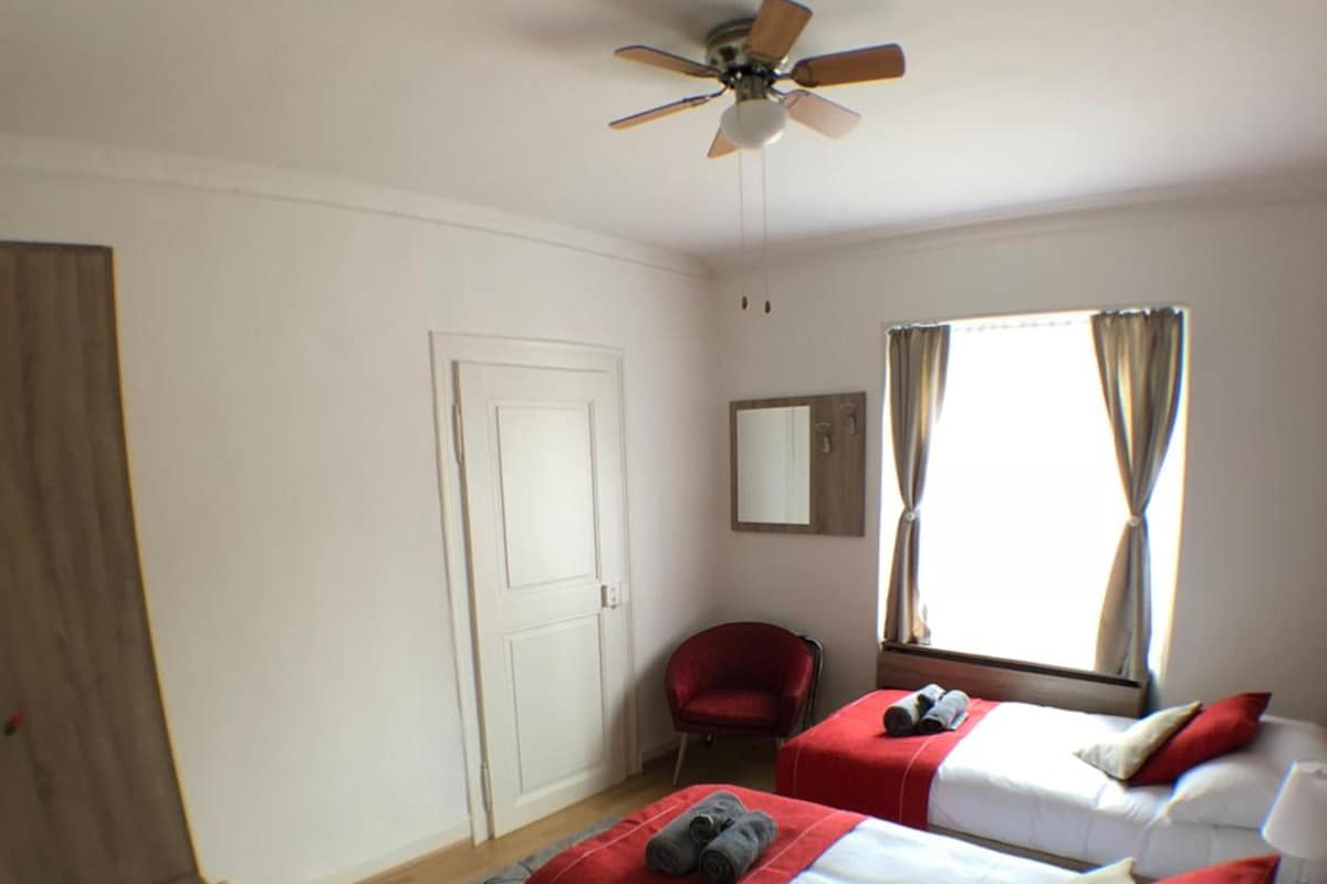 Habitación de hotel con dos camas, un escritorio, una silla, un ventilador de techo y una ventana con cortinas.