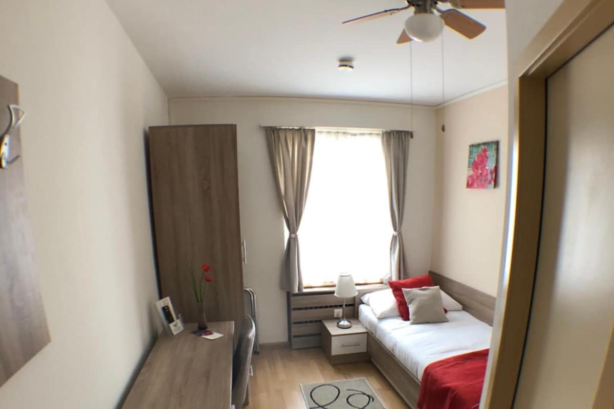 Habitación de hotel pequeña con cama, mesita de noche, escritorio y ventilador de techo.