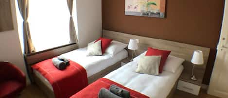 Habitación de hotel con dos camas, un ventilador de techo, una ventana con cortinas, un cuadro en la pared y una silla roja.