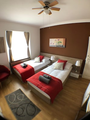 Habitación de hotel con dos camas, un ventilador de techo, una ventana con cortinas, un cuadro en la pared y una silla roja.