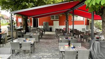 Eine Terrasse im Freien mit Tischen und Stühlen, einer roten Markise und einem Gebäude im Hintergrund.