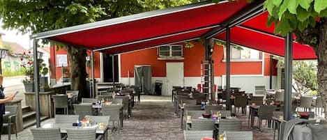An outdoor patio with tables and chairs, a red awning, and a building in the background.