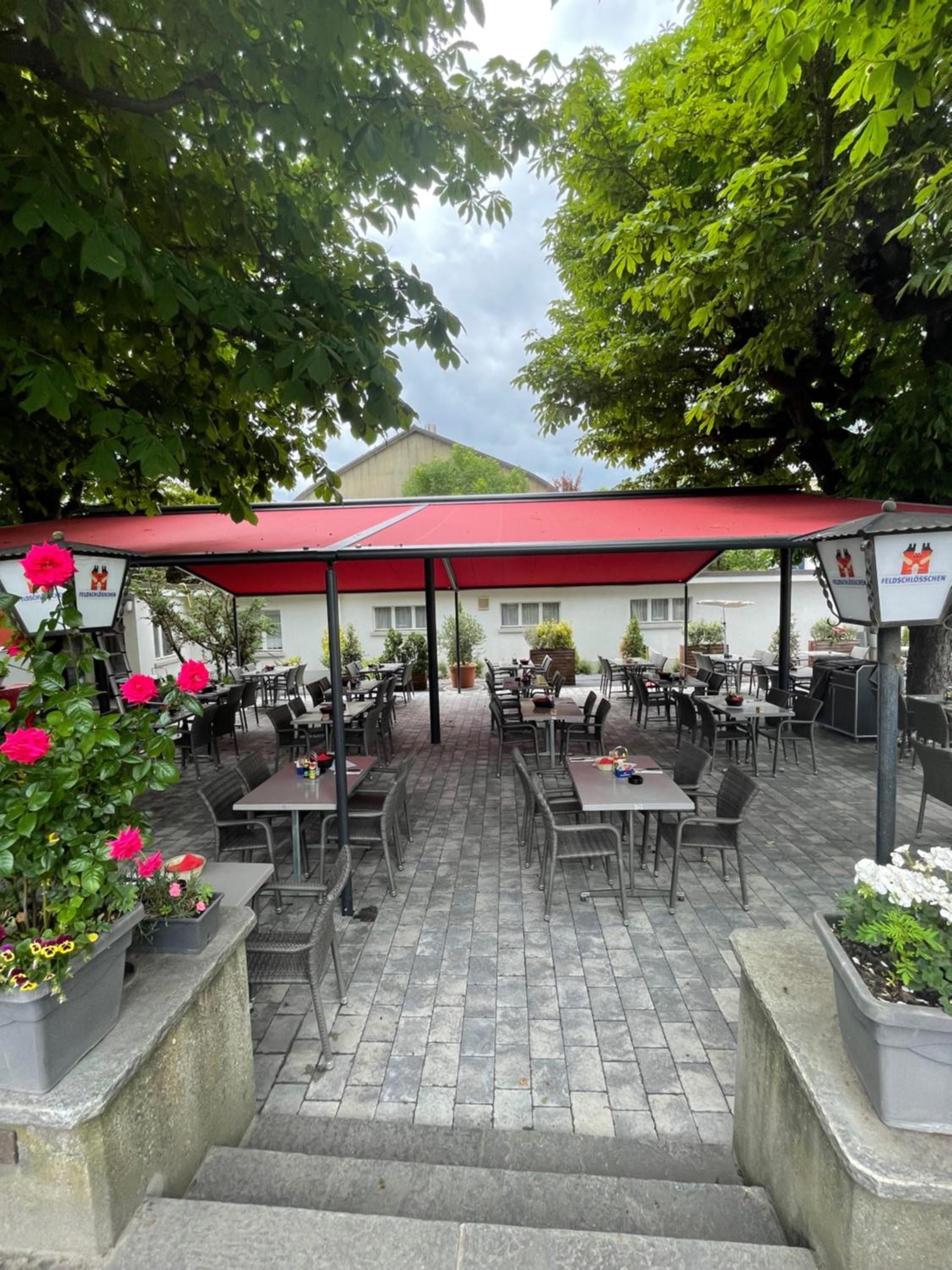 An outdoor patio with tables and chairs, a red awning, and potted plants.