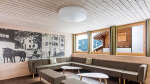 A modern interior with a wooden ceiling, a large wall mural, a sofa set, and a view through the windows. - Nova Casa Spinatscha Sedrun (Tujetsch)