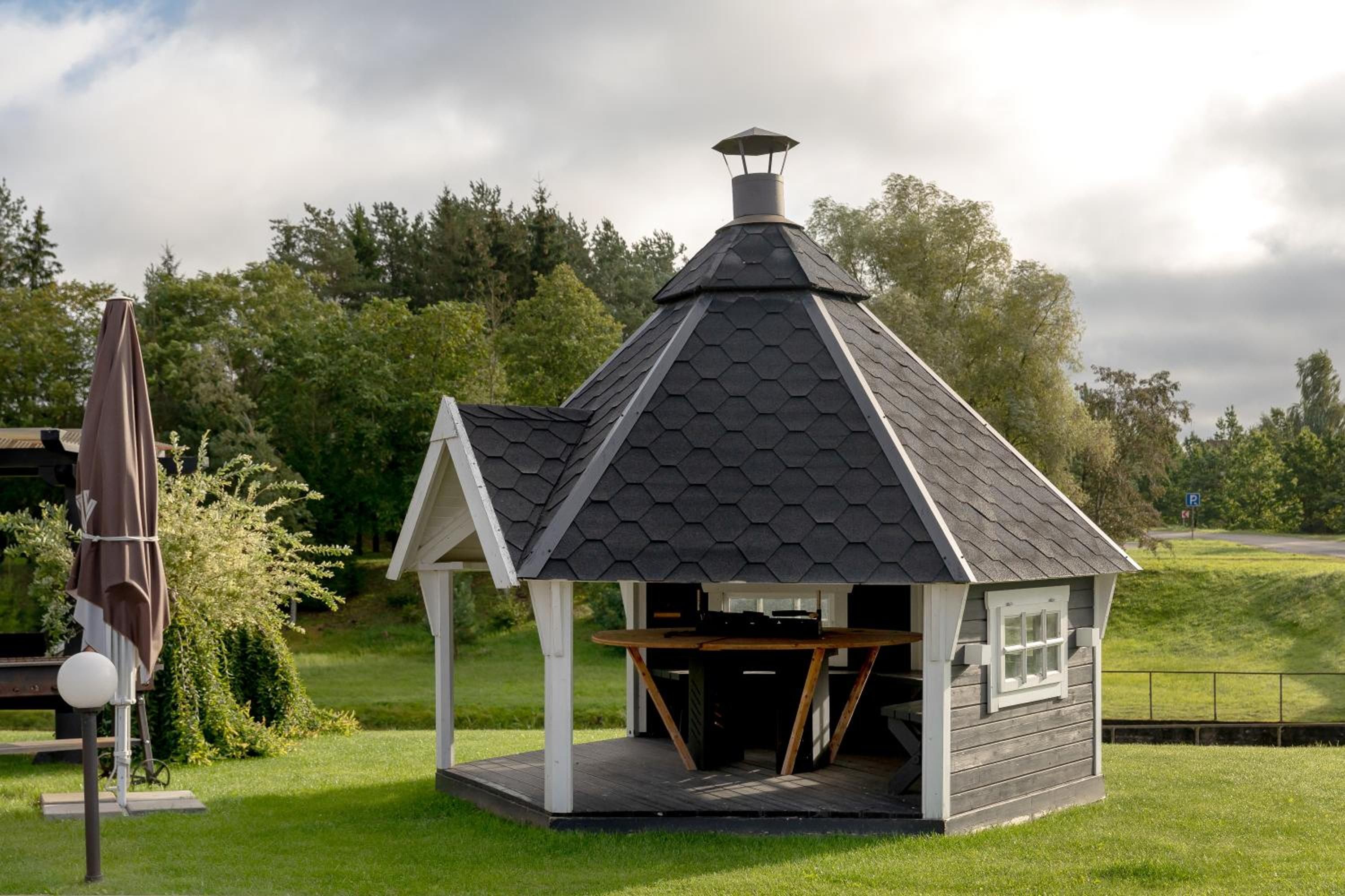 A small, hexagonal gazebo with a peaked roof and a chimney, situated on a grassy lawn with trees in the background.