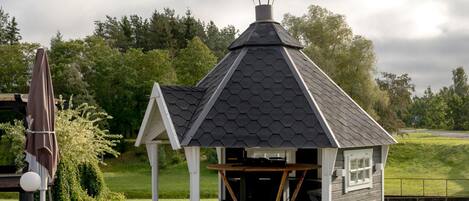 A small, hexagonal gazebo with a peaked roof and a chimney, situated on a grassy lawn with trees in the background.