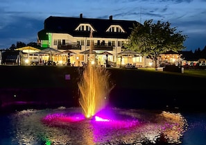 A building with a large fountain in front, surrounded by outdoor seating areas and a tree. - Butenas Hotel Tyla (Biržai)