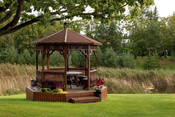 A wooden gazebo with a roof, surrounded by a grassy area, near a body of water. - Butenas Hotel Tyla (Biržai)