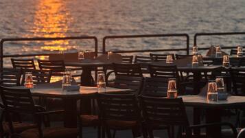 Un espace extérieur aménagé pour les repas, avec des tables et des chaises, offrant une vue sur un plan d’eau au coucher du soleil.