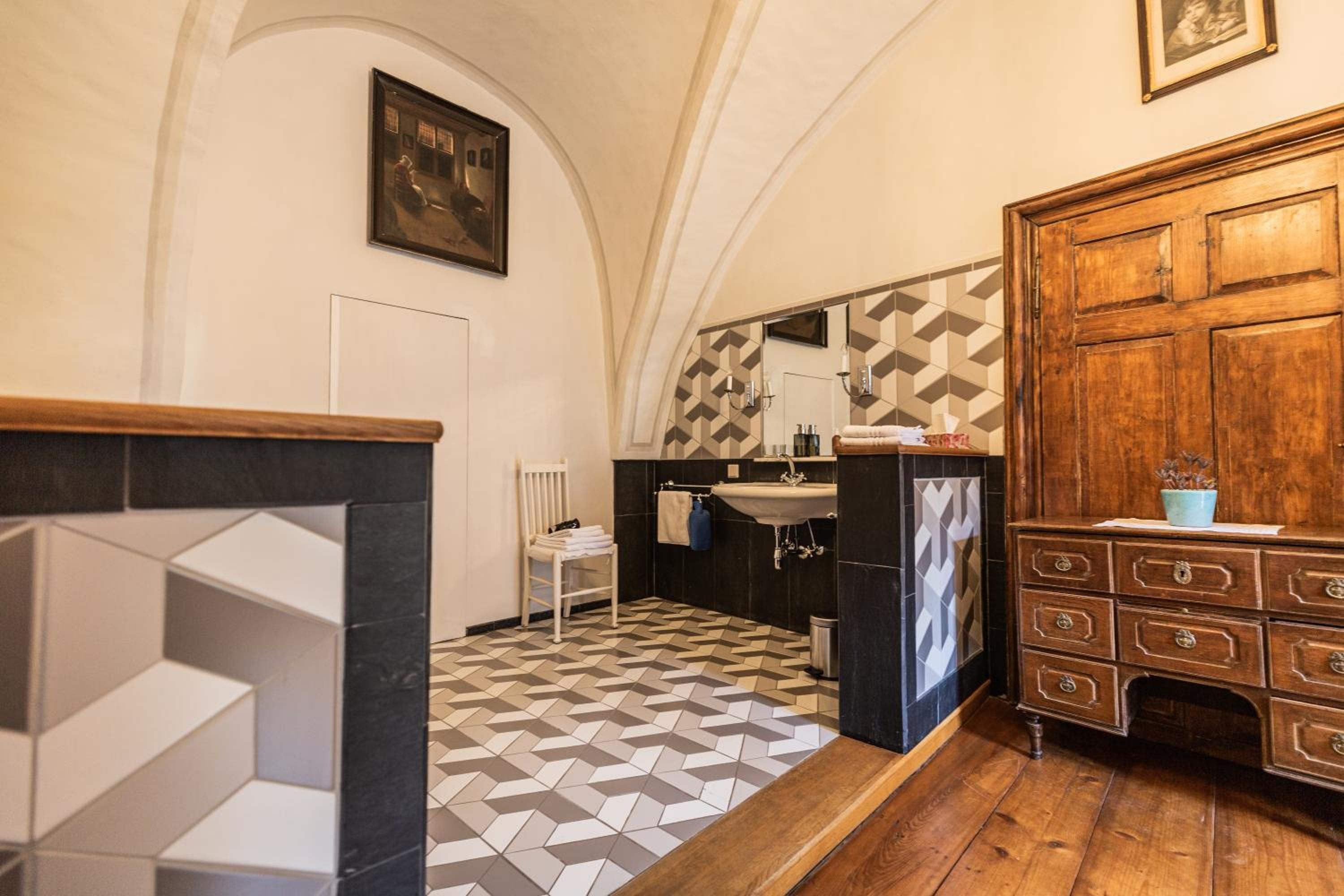 Un baño con piso de diseño geométrico, un mueble de madera con cajones, una silla blanca y un espejo sobre el lavamanos.