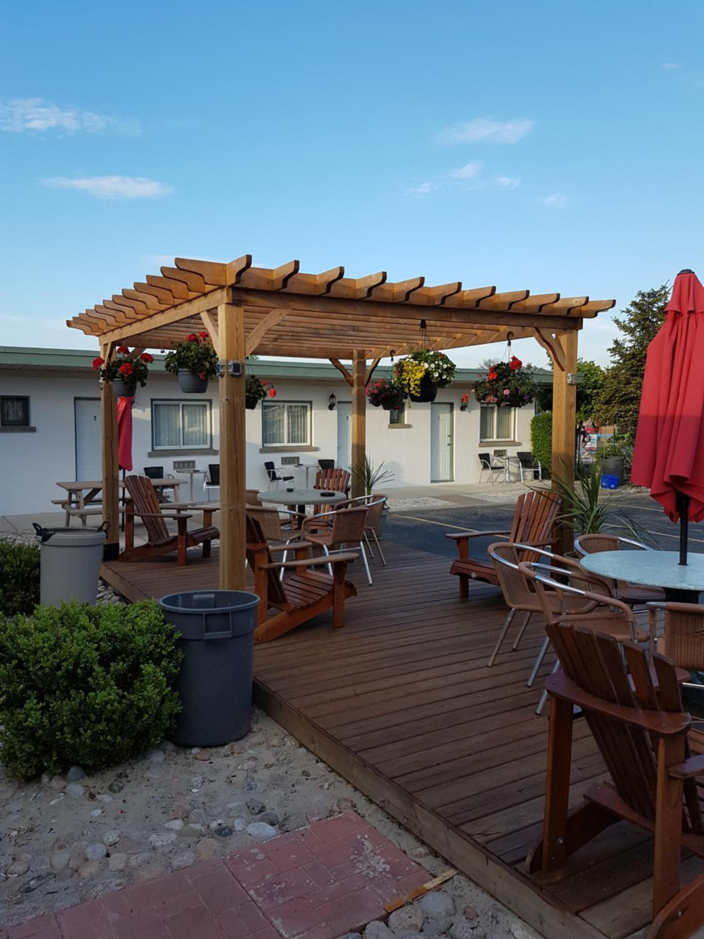 An outdoor patio area with wooden furniture, a pergola with hanging flower baskets, and a red umbrella.