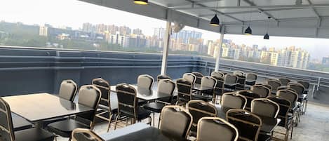 A rooftop terrace with tables and chairs, a city view, and hanging pendant lights.