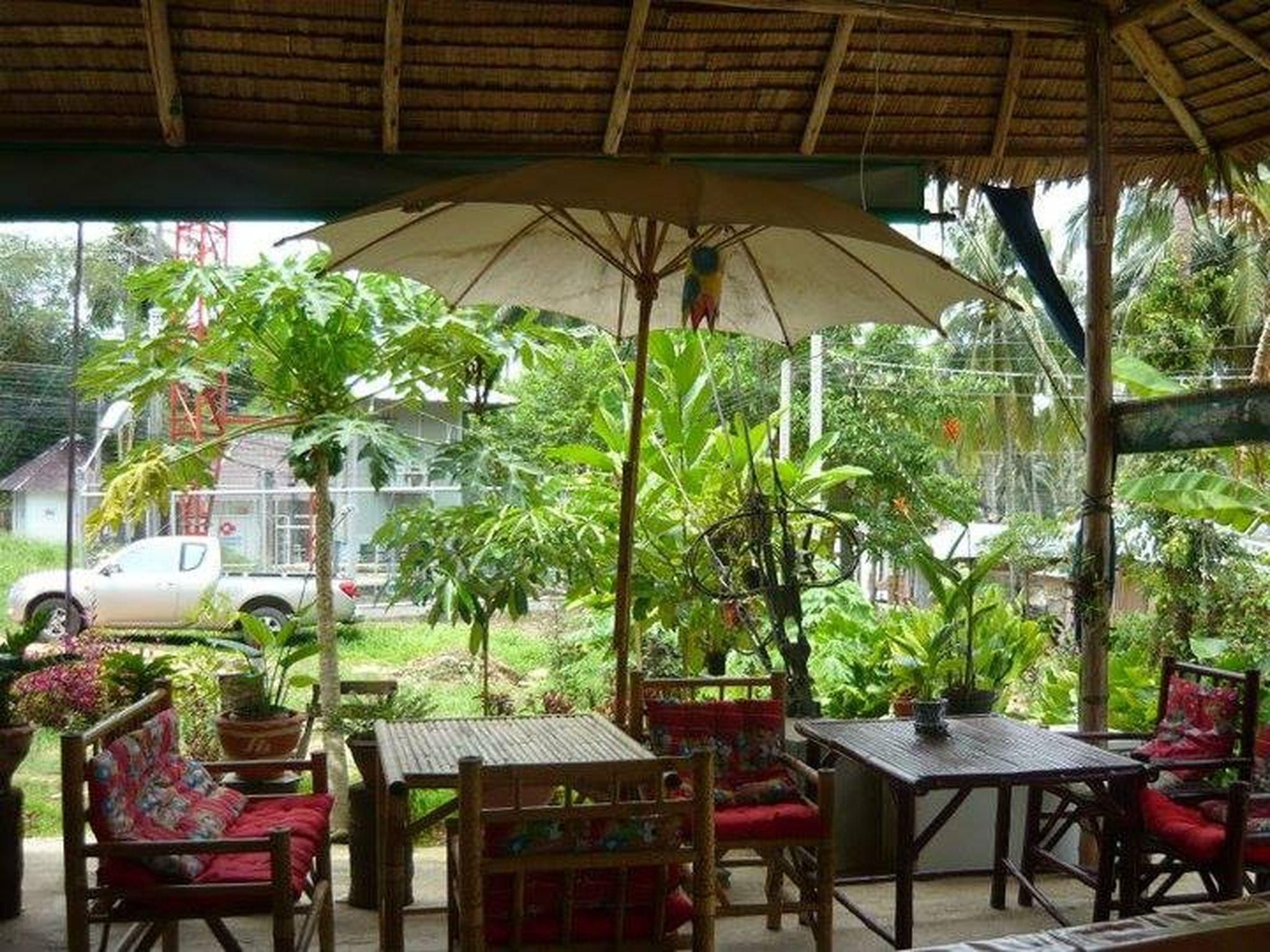 Un área de estar al aire libre con mesas y sillas, rodeada de abundante follaje y una estructura de techo de paja.