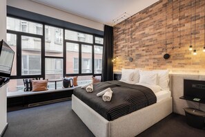 A modern hotel room with a large bed, a TV, a brick-patterned wall, and a window with a view of buildings. - Vaci Boutique Loft Suites By Bqa (Budapest)