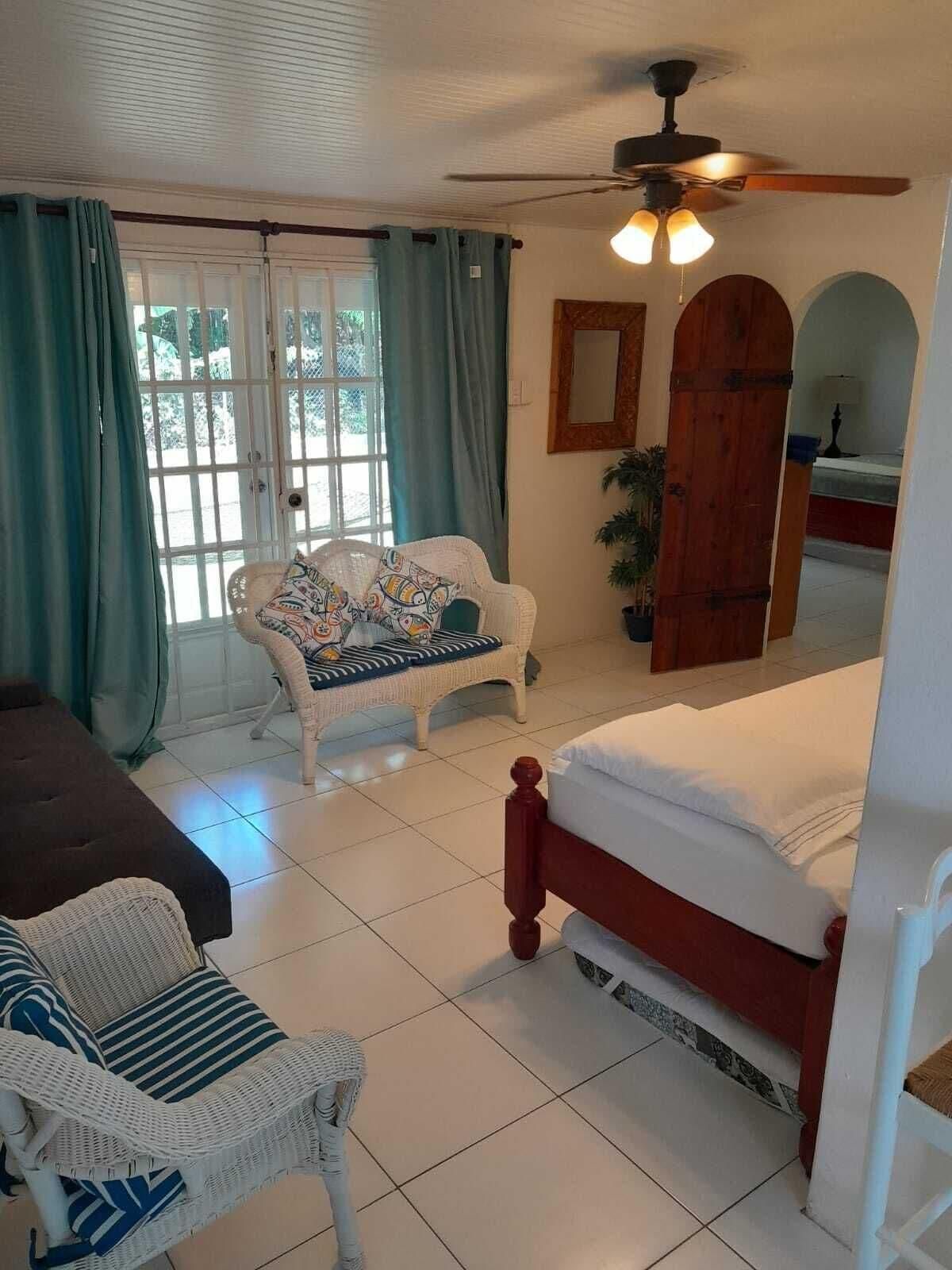 A room with a bed, a wicker chair, a sofa, a ceiling fan, and a door with a mirror.